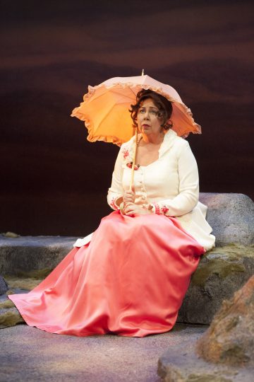 Valkoisen ja vaaleanpunaisen mekon nainen istuu kalliolla pitelemässä persikanväristä aurinkovarjoa. Hänellä on kiharat hiukset ja mietteliäs ilme, ja taustalla on dramaattinen, tumma taivas.