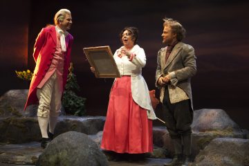 Lavalla esiintyy kolme aikakauden pukuihin pukeutunutta näyttelijää. Yksi valkoiseen toppiin ja vaaleanpunaiseen hameeseen pukeutunut nainen pitelee kehystettyä esinettä ja puhuu, kun taas kaksi vanhanaikaiseen pukuun pukeutunutta miestä seisoo molemmin puolin, kivien ja maisemien keskellä.