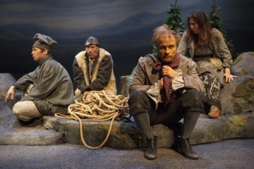 Neljä näyttelijää karuissa, vanhanaikaisissa puvuissa istuu näyttämöllä kallioisessa maastossa, ja heidän vieressään on kelautuneita köysiä ja taustalla mäntyjä, jotka viittaavat dramaattiseen tai historialliseen teatterikohtaukseen.