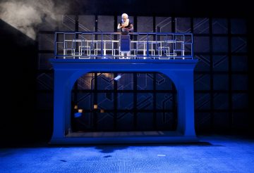 Henkilö seisoo korotetulla korokkeella, jossa on geometriset kaiteet, nojautuu eteenpäin ja katsoo alaspäin dramaattisen sinisen näyttämövalaistuksen ja taustalla olevan kevyen sumun alla.