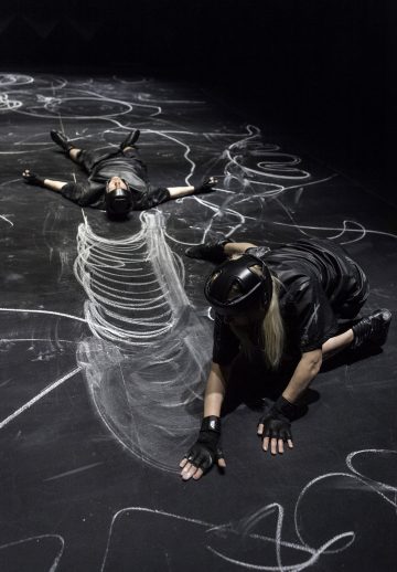 Kaksi mustiin kypäröihin ja hanskoihin pukeutunutta henkilöä on tummalla lattialla, jossa on valkoisia liitupiirroksia; toinen henkilö makaa selällään, kun taas toinen polvistuu ja näyttää ulottuvan suuren, kylkiluiden kaltaisen liitupiirroksen väliin.