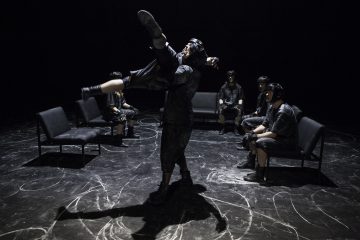 Kaksi tummiin pukuihin pukeutunutta ihmistä tekee dramaattisen hissin näyttämön keskellä, ja muut istuvat tuoleilla ympyrän sisällä. Näyttämön lattia on merkitty valkoisilla ympyräkuvioilla, ja kohtaus on hämärästi valaistu, ja siinä vallitsee teatterillinen tunnelma.