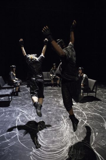 Kaksi mustiin asuihin pukeutunutta ihmistä hyppää ilmaan kädet kohollaan pimeässä huoneessa, kun taas muut samalla tavalla pukeutuneet istuvat ympyrän muotoisilla tuoleilla, ja lattialla on liitumerkintöjä.