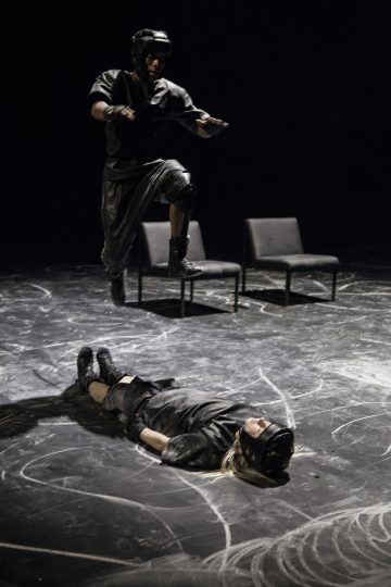 Mustiin suojavarusteisiin pukeutunut henkilö hyppää ilmaan toisen maassa makaavan henkilön yläpuolelle, jolla myös on suojavarusteet. Taustalla näkyy kaksi tyhjää tuolia liidulla merkityllä lavalla.