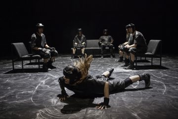 Mustiin suojavarusteisiin ja kypärään pukeutunut henkilö tekee dynaamista liikettä lattialla, kun viisi muuta samanlaisiin varusteisiin pukeutunutta henkilöä istuu tuoleilla ja sohvalla hämärästi valaistussa huoneessa ja tarkkailee tarkkaavaisesti.