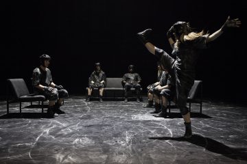 Viisi tummiin vaatteisiin ja kypärään pukeutunutta ihmistä istuu mustilla tuoleilla ympyrässä hämärästi valaistulla näyttämöllä, kun taas kuudes henkilö seisoo edessä ja potkaisee yhtä jalkaa korkealle ilmaan. Lattialla on valkoisia raitoja.