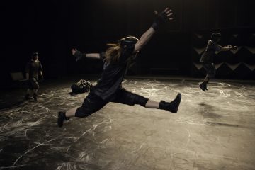 Kolme tummiin vaatteisiin ja kypärään pukeutunutta henkilöä tekee dynaamisia hyppyjä ja tanssiliikkeitä hämärästi valaistulla näyttämöllä, joka on merkitty valkoisilla liituviivoilla ja -muodoilla.