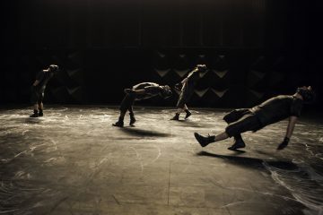 Neljä tummiin asuihin ja kypäriin pukeutunutta tanssijaa esittää dramaattisia, synkronoituja nojaavia asentoja hämärästi valaistulla näyttämöllä, jonka lattialla on liitumerkintöjä ja tumma tausta.