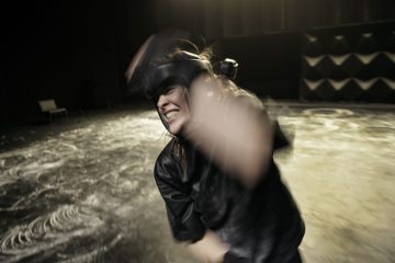 Mustiin suojavarusteisiin pukeutunut henkilö on keskellä toimintaa ja heiluttaa käsivarttaan voimallisesti epäselvässä liikkeessä, päättäväinen ilme kasvoillaan, hämärässä, tilavassa huoneessa.