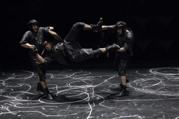 Kolme mustiin urheiluvarusteisiin ja kypärään pukeutunutta henkilöä esiintyy tummalla näyttämöllä, jossa on valkoisia liituviivoja; kaksi pitää kolmatta henkilöä vaakatasossa ilmassa.