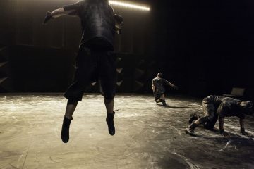 Kolme tummiin vaatteisiin pukeutunutta tanssijaa esiintyy hämärästi valaistulla näyttämöllä. Yksi on ilmassa, toinen on kyyryssä ja kolmas polvillaan, mikä viittaa dynaamiseen, urheilulliseen liikkeeseen.