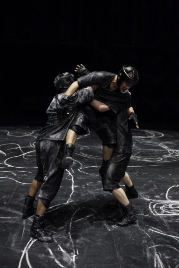 Kaksi mustiin suojavarusteisiin ja kypärään pukeutunutta henkilöä kamppailee liidulla merkityllä näyttämöllä kesken toiminnan, ja toinen henkilö nostaa jalkaansa painin aikana. Tausta on tumma ja kohtaus vaikuttaa intensiiviseltä.