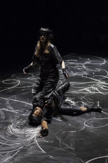 Kaksi mustiin suojavarusteisiin ja kypäriin pukeutunutta ihmistä on liitupeitteisellä lattialla; toinen seisoo toisen päällä, joka makaa maassa. Kohtaus viittaa lavastettuun tai koreografioituun taisteluun tai esitykseen.