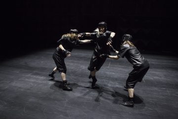 Kolme mustiin urheiluasuihin ja kypärään pukeutunutta ihmistä esittää ilmaisuvoimaista, dynaamista tanssia tai teatraalista liikettä pimeällä, tyhjällä näyttämöllä, ja kaksi heistä kurottautuu kohti keskellä olevaa kolmatta.