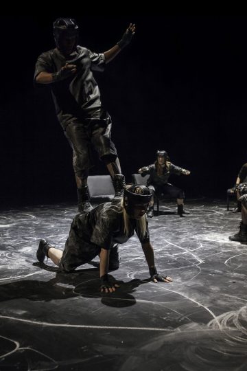 Kaksi kypärä- ja suojavarusteisiin pukeutunutta henkilöä esiintyy liidulla merkityllä lavalla; toinen kyykistyy nelinkontin ja toinen seisoo selällään. Kaksi muuta esiintyjää istuu taustalla, kädet ojennettuina tasapainon vuoksi.