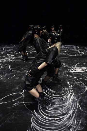 Mustiin kypäröihin ja asuihin pukeutuneet esiintyjät nojaavat pareittain toisiaan vasten tummalla näyttämöllä, joka on peitetty valkoisella liidulla tai maalilla, ja luovat niiden alle dynaamisia kuvioita.