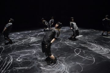 Seitsemän tanssijaa, joilla on kypärät ja tummat vaatteet, esiintyvät valkoisten liituviivojen ja tahrojen peittämällä näyttämöllä, ja jokainen heistä ottaa dynaamisia, nojailevia asentoja dramaattisessa valaistuksessa.