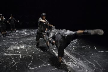 Neljä ihmistä pehmustetuissa varusteissa ja kypärissä esiintyy valkoisilla liitumerkinnöillä peitetyllä lavalla. Yksi henkilö on keskellä ilmaa ja suorittaa potkun, kun taas toinen seisoo vastakkain, hansikkaat kädet kohotettuna estämään tai ottamaan potkun kiinni.