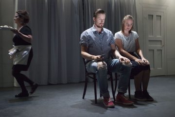 Mies ja nainen istuvat vierekkäin tuoleilla valokeilan alla ja katsovat vakavina eteenpäin. Taustalla kulkee tarjoilija tarjottimensa kanssa, ja heidän takanaan näkyy harmaita verhoja ja ovi.