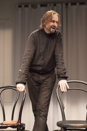 Olkapäiden pituiset hiukset ja partainen, kokonaan mustiin pukeutunut mies seisoo nojaamassa kahteen mustaan tuoliin valkoisten verhojen edessä, katsoo sivulle ja hymyilee.