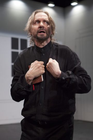 Pitkätukkainen, parrakas, mustiin pukeutunut mies puristaa pientä kirjaa rintaansa ja katsoo ylöspäin intensiivinen, tunteikas ilme. Hän seisoo kirkkaiden näyttämövalojen alla minimalistisessa huoneessa.