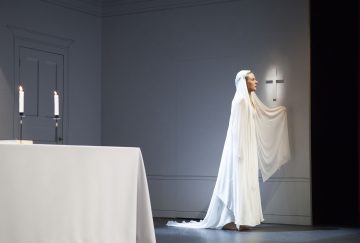 Valkoiseen pitkään pukuun ja huntuun pukeutunut nainen seisoo lavalla valaistun ristin seinän vieressä ja pitää huntua. Etualalla valkoisella peitetyllä pöydällä on kaksi palavaa kynttilää. Lavastus on minimalistinen ja dramaattinen.