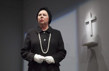 Mustaan asuun pukeutunut nainen, jolla on musta turbaani, valkoiset hansikkaat ja pitkä helmikaulakoru, seisoo kädet ristissä. Hänen vieressään seinälle on kiinnitetty valkoinen risti. Asetelma vaikuttaa synkältä ja muodolliselta.