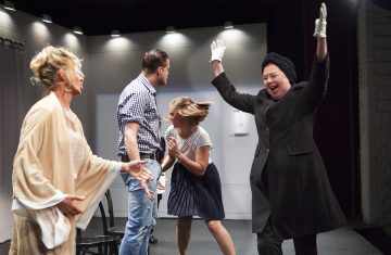 Neljä näyttelijää näyttämöllä, yksi mustapukuinen nainen, jolla on kohotetut kädet ja valkoiset hanskat, vaikuttaa innostuneelta, kun taas muut reagoivat tarmokkaasti; kohtaus näyttää dramaattiselta ja ilmeikkäältä teatterivalaistuksen ansiosta.