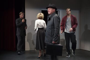 Lavalla seisoo neljä ihmistä: mustaan takkiin ja hattuun pukeutunut mies pitelee salkkua ja katsoo eteenpäin, kun punatakkinen mies ja toinen mustapukuinen mies puhuvat; vintage-asuun pukeutunut nainen seisoo selkä kameraan päin.