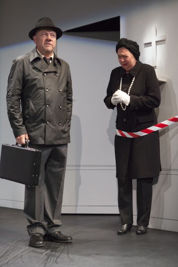 Mustaan trenssitakkiin ja hattuun pukeutunut mies pitelee salkkua, kun taas mustaan hautajaisasuun pukeutunut nainen seisoo punavalkoisen nauhan takana ja vaikuttaa järkyttyneeltä valkoisen seinän ja ristin lähellä.