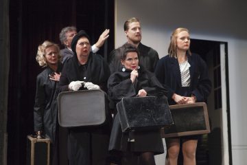 Lavalla tummiin, virallisiin vaatteisiin pukeutunut kuuden näyttelijän ryhmä pitää matkalaukkuja kädessään ja katsoo vakavin ja huolestunein ilmein eteenpäin. Lavastus on minimaalinen, ja taustalla on oviaukko.