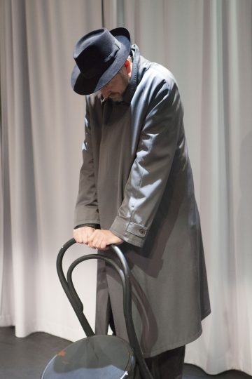 Tummiin takkeihin ja hattuihin pukeutunut mies seisoo pää alaspäin kumarassa, molemmat kädet tuolin selkänojalla, valkoisten verhojen edessä.