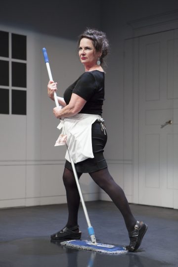 Mustaan mekkoon, esiliinaan ja sukkahousuihin pukeutunut nainen seisoo luottavaisesti moppaamassa lattiaa lavasteessa, jossa on valkoiset seinät ja suljettu ovi hänen takanaan. Hänellä on siivoustarvikkeita esiliinassaan ja mustat kengät jalassaan.