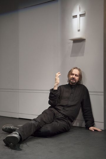 Pitkätukkainen, parrakas, mustiin pukeutunut mies istuu lattialla seinää vasten, ja hänen yläpuolellaan on valkoinen risti. Hän elehtii yhdellä kädellä ja vaikuttaa mietteliäältä tai uupuneelta.