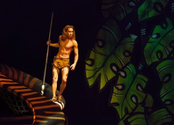 Tarzaniksi pukeutunut henkilö, jolla on lannevaate ja keihäs kädessään, seisoo suuren, raidallisen, viidakon oksaa muistuttavan rakennelman päällä. Taustalla näkyy värikkäitä, ylisuuria vihreitä lehtiä.