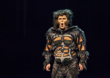 Henkilö, jolla on teatterimainen kissa-asu, jossa on turkki, dramaattinen meikki ja leikattuja kuvioita rinnassa, seisoo lavalla, suu auki kuin laulaisi, tummaa taustaa vasten.