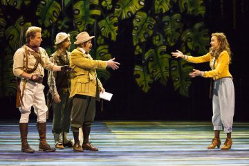 Neljä näyttelijää värikkäissä, vintage-tyylisissä puvuissa seisoo lavalla, jonka taustalla on vehreä viidakko. Kolme miestä ojentaa hymyilevän naisen kohti ojennetut kädet, ikään kuin tervehtien tai toivottaen hänet tervetulleeksi.