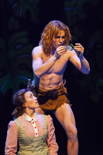 Karkeaan lannevaatteeseen pukeutunut mies tutkii tarkkaan vihreää esinettä, kun raidalliseen puseroon ja liiviin pukeutunut nainen katsoo häntä yllättyneenä; he näyttävät olevan teatteriesityksen näyttämöllä.