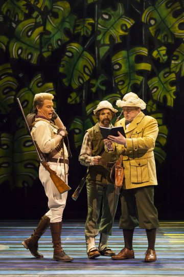 Lavalla seisoo kolme tutkimusmatkailija-asuihin pukeutunutta näyttelijää. Yhdellä on kädessään kivääri, toisella verkko, ja kolmas lukee leikepöydästä. Taustalla on suuria, tyyliteltyjä vihreitä lehtiä, jotka viittaavat viidakkoympäristöön.