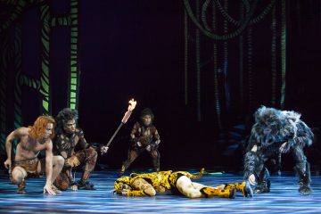 Viisi esiintyjää eläimistä inspiroiduissa puvuissa toimii lavalla; yksi makaa liikkumatta lattialla, kun muut kyyristelevät tai nojaavat heitä kohti, ja yksi pitää taskulamppua. Taustalla on viidakkomaisia koristeita.