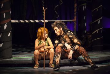 Vihreään pukuun pukeutunut lapsi, jolla on sauva kädessään, istuu tiikerin asuun ja meikkiin pukeutuneen aikuisen vieressä lavalla, jossa on viidakkoaiheista rekvisiittaa ja hämärä valaistus. Molemmat näyttävät näyttelevän teatteriesityksessä.