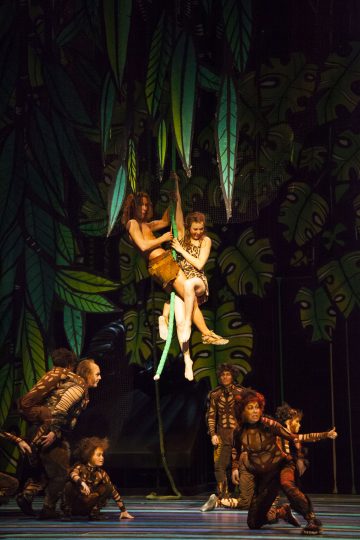 Viidakkoaiheisiin pukuihin pukeutuneet esiintyjät ovat lavalla. Kaksi ihmistä roikkuu köynnöksen varassa muiden yläpuolella, ja taustalla on suuria vihreitä lehtiä. Kohtaus tuo mieleen vilkkaan, teatraalisen viidakkoympäristön.