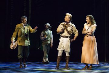 Neljä näyttelijää aikakauden puvuissa seisoo näyttämöllä dramaattisen valaistuksen alla. Yksi mies elehtii vilkkaasti, toinen seisoo kädet lanteilla, kolmas pitelee kivääriä taustalla, ja pitkämekkoon pukeutunut nainen katselee tilannetta kädet ristissä.