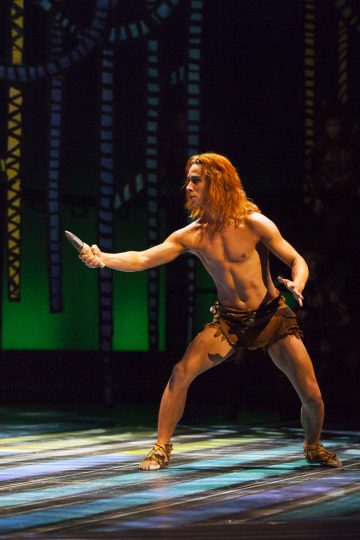 Paidaton pitkätukkainen mies, jolla on ruskea lannevaate ja sandaalit, seisoo dramaattisessa poseerauksessa veitsi kädessään lavalla, jossa on värikäs, abstrakti tausta.