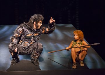 Mies karussa, eläimistä inspiroituneessa puvussa kyyristelee ja elehtii puhuessaan lapselle, joka on pukeutunut nuoreksi luolamökkiläiseksi ja pitää keihästä kädessään, molemmat näyttämöllä, jossa on dramaattinen valaistus.