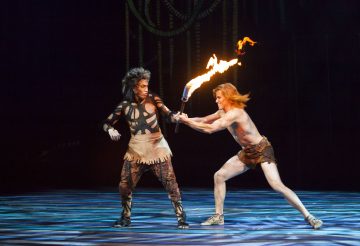 Lavalla esiintyy kaksi heimopukuista esiintyjää, joista toinen pitää kädessään liekehtivää soihtua, kun toinen yrittää tarttua siihen. Kohtaus on dynaaminen ja dramaattinen, ja siinä on elävä valaistus ja tumma tausta.