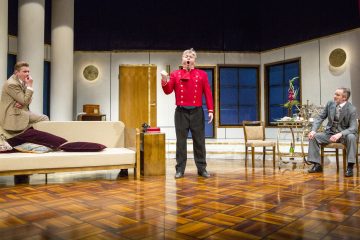 Kolme miestä esiintyy näyttämönäytelmässä, joka sijoittuu tyylikkääseen huoneeseen, jossa on valkoiset pylväät ja modernit huonekalut. Yksi punapukuinen mies seisoo keskellä ja elehtii vilkkaasti, kun taas kaksi muuta miestä istuvat ja katselevat häntä tarkkaavaisesti.