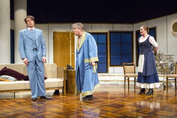 Kolme näyttelijää esiintyy olohuoneen näköisessä lavastuksessa. Yksi siniseen pukuun pukeutunut mies seisoo jäykästi, toinen siniseen kaapuun pukeutunut mies vihoittelee häntä kohti, kun taas mekkoon pukeutunut nainen seisoo sivussa ja vaikuttaa yllättyneeltä.