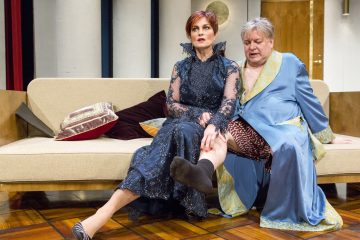 Kaksi ihmistä istuu tyylikkäissä, vintage-tyylisissä kylpytakeissa beigen värisellä sohvalla. Nainen laivastonsinisessä pitsimekossa katsoo vakavana eteenpäin, kun taas siniseen kaapuun ja eläinkuvioisiin housuihin pukeutunut mies istuu hänen takanaan pitelemässä hänen jalkaansa.