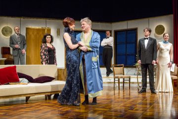 Seitsemän näyttelijää tyylikkäissä iltapuvuissa ja kaapuissaan esiintyy tyylikästä olohuonetta muistuttavalla lavasteella, jossa kaksi keskeistä hahmoa keskustelee muiden katsellessa, mikä viittaa dramaattiseen tai komedialliseen teatterikohtaukseen.